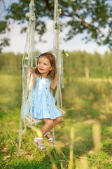 A nice little girl in a blue dress is swinging on a swing decorated with ribbons and flowers. The child looks away smiling sweetly