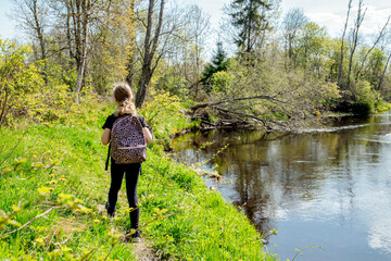 Back view of 9 year old girl hiking, walking alone in the forest by the river with a backpack in early spring. Beautiful idyllic nature.