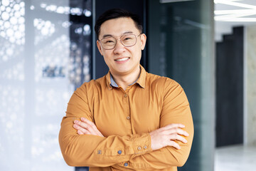 Portrait of happy and successful asian businessman man in glasses smiling and looking at camera, manager business owner with crossed arms near window working inside modern office