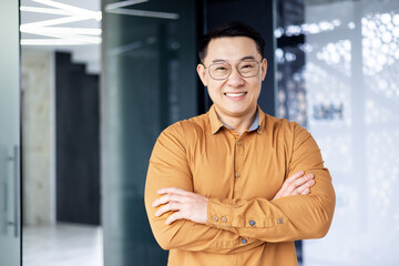 Obraz premium Portrait of successful asian businessman, man inside office wearing shirt and glasses smiling and looking at camera, investor broker with crossed arms near window.