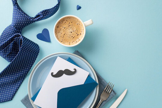 Beautifully arranged Father's Day table setting with top view flat lay of dishes, envelope, postcard with mustaches, napkin, tie, coffee on a pastel blue background with empty space for text or ad