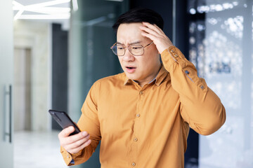 Upset asian man in office at work reading bad news from phone online, businessman in shirt near...