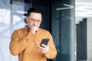 Asian businessman inside office building upset and sad reading bad news on mobile phone, business...