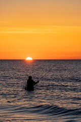 Naklejka na ściany i meble Silhouette eines Angler in wasserdichten Hosen welcher vor einem traumhaften orangen Sonnenuntergang in der Ostsee angelt / fischt . Er steht mitten im Wasser der Ostsee.