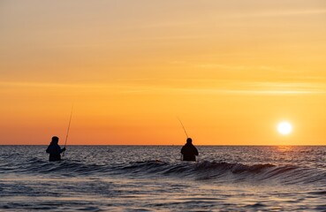 Die Silhouetten von zwei  Anglern in wasserdichten Hosen angelen vor einem traumhaften orangen...