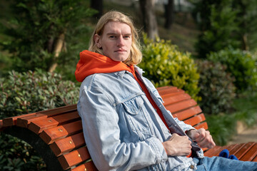 Concentrated scandinavian hipster guy sitting on wooden bench looking at camera on sunny day....