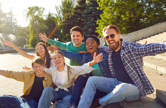 Bunch of happy multiracial friends having fun outdoors. Diverse group of cheerful joyful young people sitting on stone steps on beautiful small city square, looking at camera and smiling all together