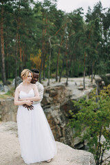 Fototapeta na wymiar Happy interracial couple newlyweds stands on rock and embraces against background of forest and canyon.