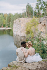 Happy interracial couple newlyweds sits on rock and embraces against beautiful background of lake, forest and canyon.