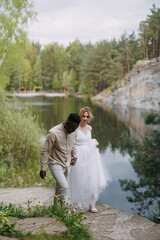 Happy interracial couple newlyweds holds each other hands and walks against background of lake, rocks and forest.