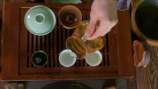 Hand of tea master pouring fresh green oolong brewing tea from glass pot to ceramic tea bowls on wooden table chaban, top view. Traditional chinese tea ceremony. Prepare serve hot beverage drink.