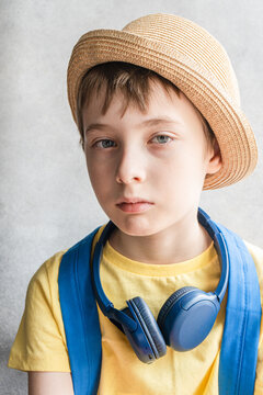 Portrait of serious kid with blue eyes and brown hair wearing yellow t-shirt and hat with blue bag pack and headphones is looking at camera