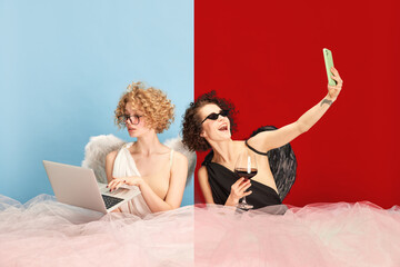Young woman in image of angel and demon sitting at table and using gadgets against blue red...