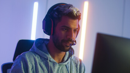 Attractive Middle East man putting on headphone playing computer SPF or MMORPG game looking at screen focus on match commanding team touching microphone. Handsome young male sit in gaming room