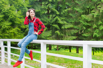 Happy young woman in blue denim overalls and red shirt sits on wooden fence