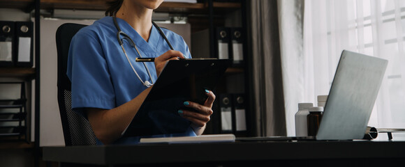 Serious female doctor using laptop and writing notes in medical journal sitting at desk. Young woman professional medic physician wearing white coat and stethoscope working on computer at workplace.