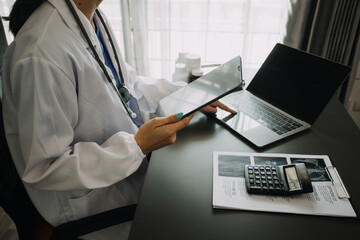 Serious female doctor using laptop and writing notes in medical journal sitting at desk. Young woman professional medic physician wearing white coat and stethoscope working on computer at workplace.