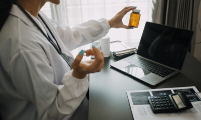 Serious female doctor using laptop and writing notes in medical journal sitting at desk. Young woman professional medic physician wearing white coat and stethoscope working on computer at workplace.