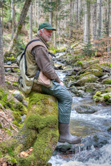An hiker is sitting on a tree stump in the forest above a mountain stream. He exudes a deep sense...