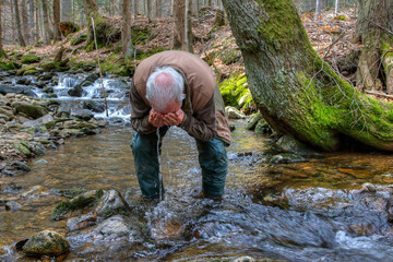 In the midst of untouched wilderness, a hiker refreshes his face in a crystal clear mountain stream with the cold water surrounded by forest and the calm murmur of the flowing creek.