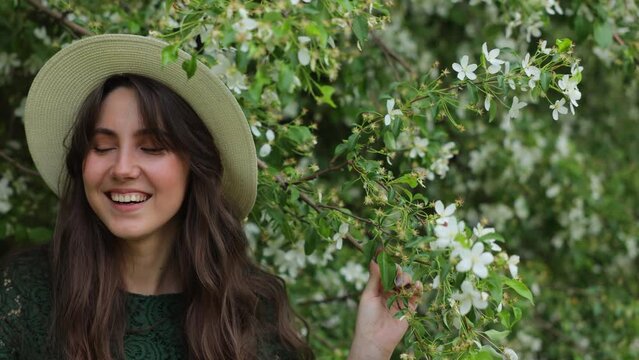 beautiful and joyful brunette woman stands in front of a blossoming apple tree and shakes it. Falling white petals from the blossoming fruit trees. Joy and happiness. The concept of spring and blossom