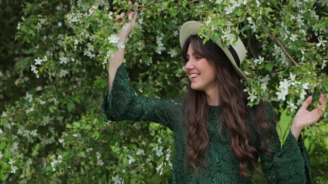 beautiful and joyful brunette woman stands in front of a blossoming apple tree and shakes it. Falling white petals from the blossoming fruit trees. Joy and happiness. The concept of spring and blossom