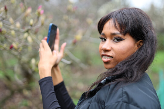 Cheerful young african girl with phone takes pictures of spring flowers in the park.