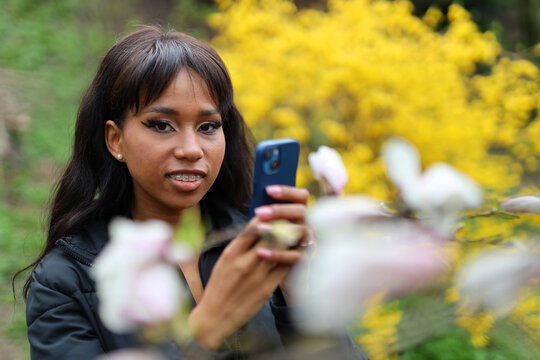 An African-American woman in a spring park photographs magnolia flowers on her phone.