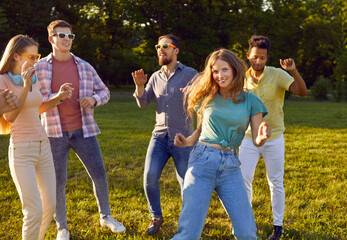Group of friends meet up and have fun at outdoor party in summer park. Happy cheerful carefree young diverse people in comfortable casual clothes dancing on fresh green lawn in city park