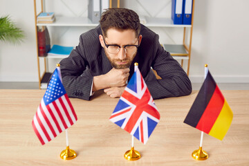 Pensive businessman sitting at desk looking at flags of different countries. Young man in formal...