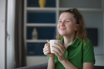 Happy young woman drinking a cup of tea. Dreaming girl sitting in living room with cup of hot coffee enjoying. Pretty woman home.