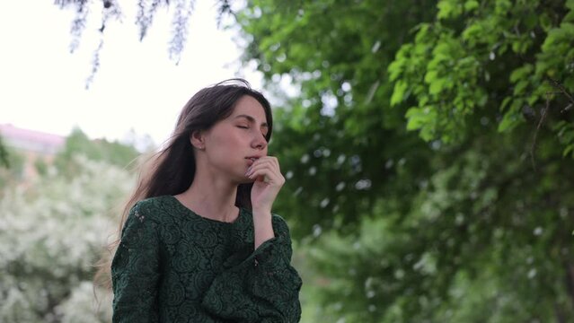 A beautiful and joyful brunette woman in a green dress stands in front of a green garden. the petals of blossoming fruit trees fall in the background. 