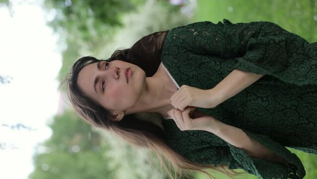 A beautiful and joyful brunette woman in a green dress stands in front of a green garden. the petals of blossoming fruit trees fall in the background. Vertical video
