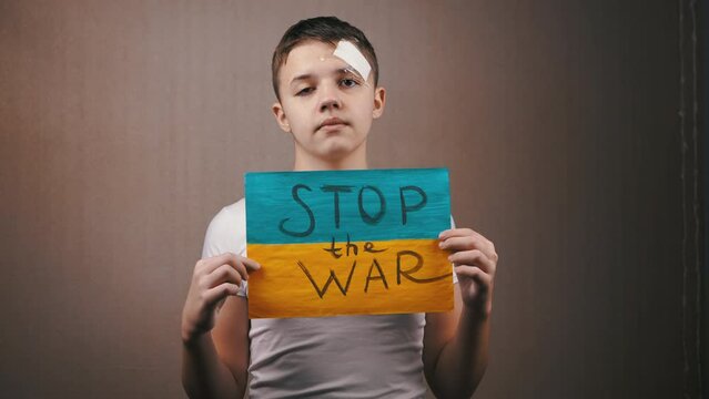 Boy Holding a Poster of the Ukrainian Flag with Inscription Stop the War. Teenage protester with tears in his eyes calls for an end to the war in Ukraine. Indoors. Grey background. Blue yellow flag.