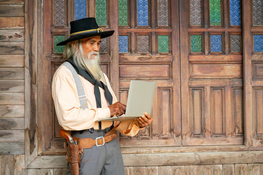 Senior Asian cowboy stand in front of wooden building and use laptop alone in concept of technology for senior people in daily life.