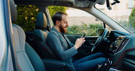 Bearded young man sitting in car and typing online message or reading news on cellphone. Male sitting in modern vehicle and using on smartphone.