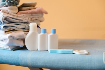 Obraz na płótnie Canvas White bottles and jars of organic natural cosmetic product for newborn baby: body oil, milk or lotion with mockup, on ironing board nearby a pile of clean laundered ironed clothes and nail scissors.
