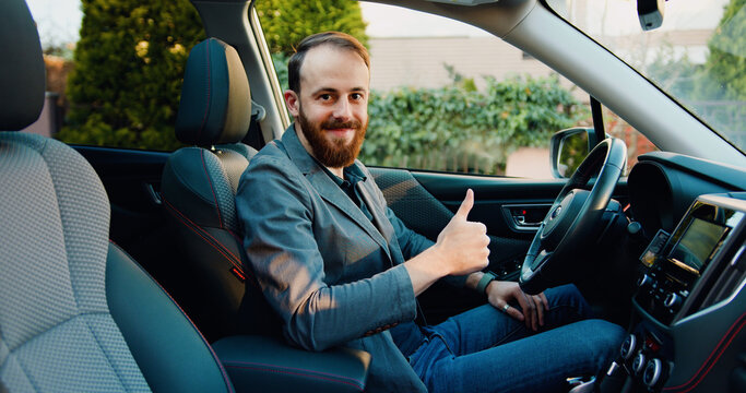 Portrait of handsome young adult man showing thumbs up sitting inside modern car looking at camera.