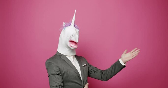 Businessman in unicorn mask gesturing in pink studio. Unrecognizable male in elegant suit and funny unicorn mask giving promotion and showing thumb up gesture of approval against pink background