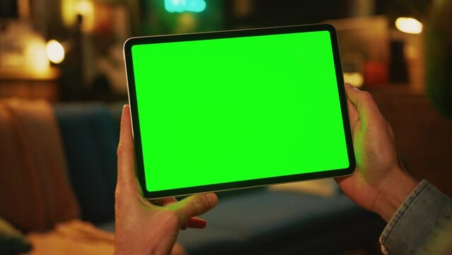 Close up hands man holding and tapping on a tablet computer with a horizontal green screen. Mock up. Sofa on background
