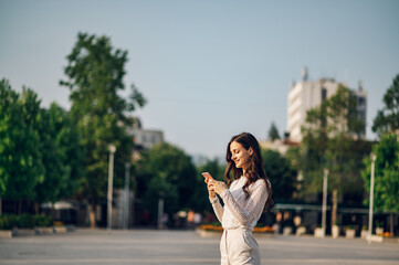 A cheerful young woman is standing on a city square downtown and texting messages on the phone.