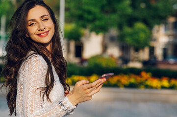 A cute fashionable young woman is standing on a city square with her phone and smiling at the camera on a sunny summer day.