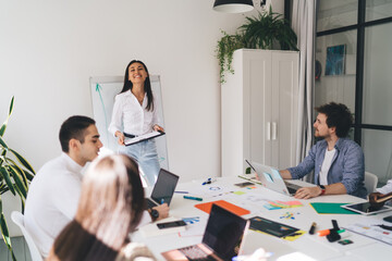 Cheerful female speaker standing in conference room presenting project to diverse coworkers