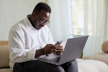 Handsome Black African American Man Working on Laptop Computer while Sitting on a Sofa in Cozy Living Room. Freelancer Working From Home. Browsing Internet, Using Social Networks, Having Fun in Flat.