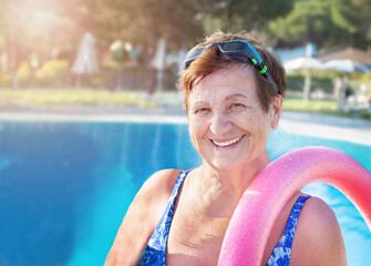 Active senior (elderly) woman (over age of 50) with swim noodles near swimming pool smiling with...