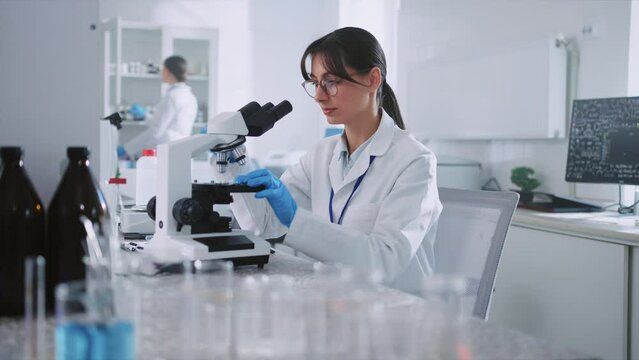Footage of good-looking young woman in gloves using equipment and looking under microscope while working in modern laboratory. Portrait of Caucasian girl in white lab coat sitting at workplace