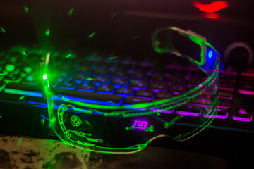 Glowing futuristic glasses with transparent glass lie on a multi-colored keyboard with glare	
