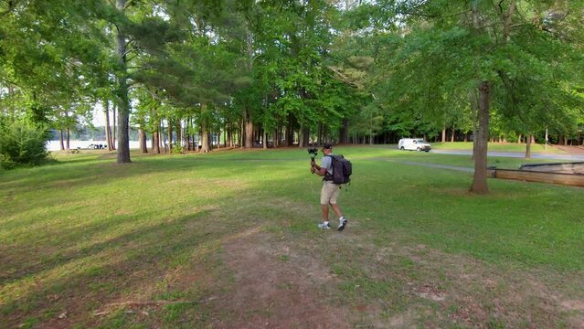 footage of an African American man wearing a backpack, filming on the banks of a lake with lush green trees and grass at sunset at Proctor Landing Park at lake Acworth in Acworth Georgia USA