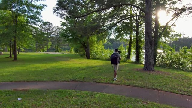 footage of an African American man wearing a backpack, filming on the banks of a lake with lush green trees and grass at sunset at Proctor Landing Park at lake Acworth in Acworth Georgia USA