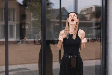 Fototapeta premium Young excited confident proud business woman winner wearing black outfit standing on street, raising hands, feeling power, motivation, energy, celebrating career financial success in big city outdoors